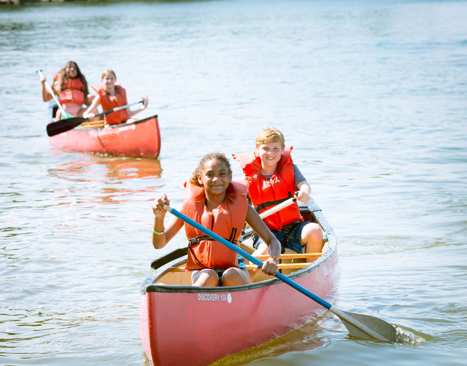 Children canoeing on a lake, wearing orange life vests. Two kids paddle a red canoe in the foreground while another canoe with two more children follows behind them. The water is calm and reflects the bright summer day.