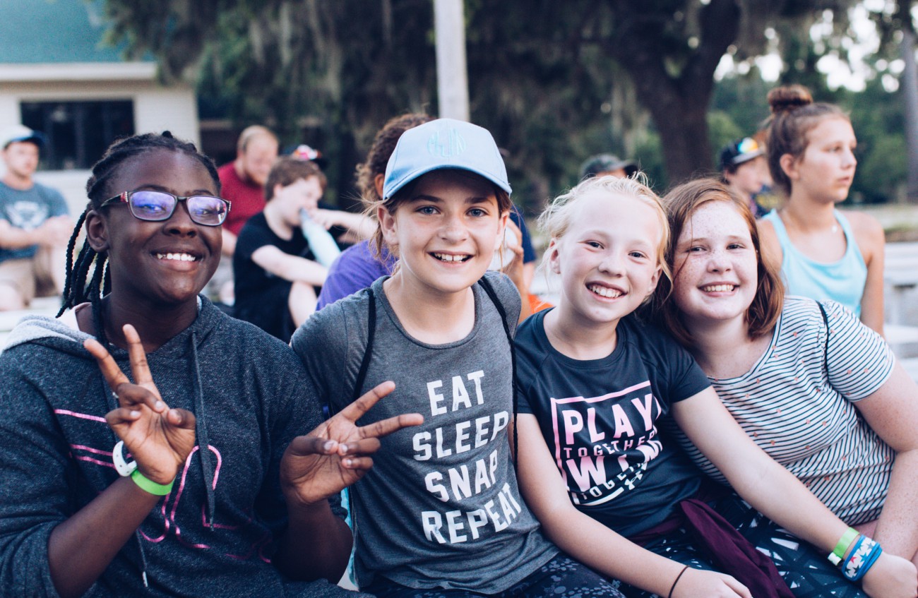 Four smiling girls at an outdoor camp showing peace signs. They wear casual t-shirts with phrases like 'EAT SLEEP SNAP REPEAT' and 'PLAY,' and some have colorful wristbands. Other campers and trees are visible in the background.