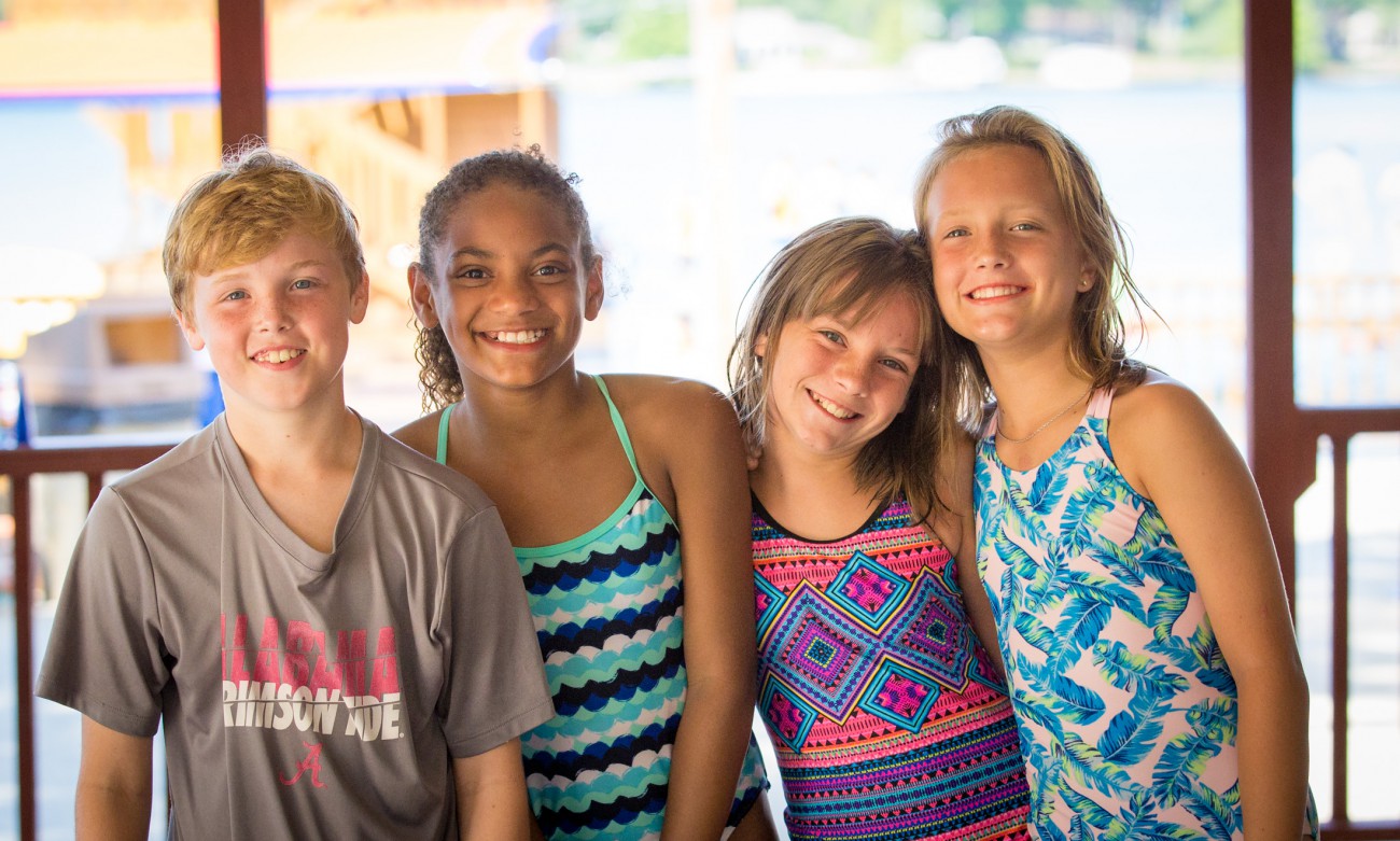 Four smiling children standing together in summer attire. From left to right: a boy in a gray Alabama t-shirt, a girl in a teal patterned swimsuit, a girl in a colorful geometric print swimsuit, and a girl in a blue tropical print outfit. They appear to be at a summer camp or resort with water visible in the background.