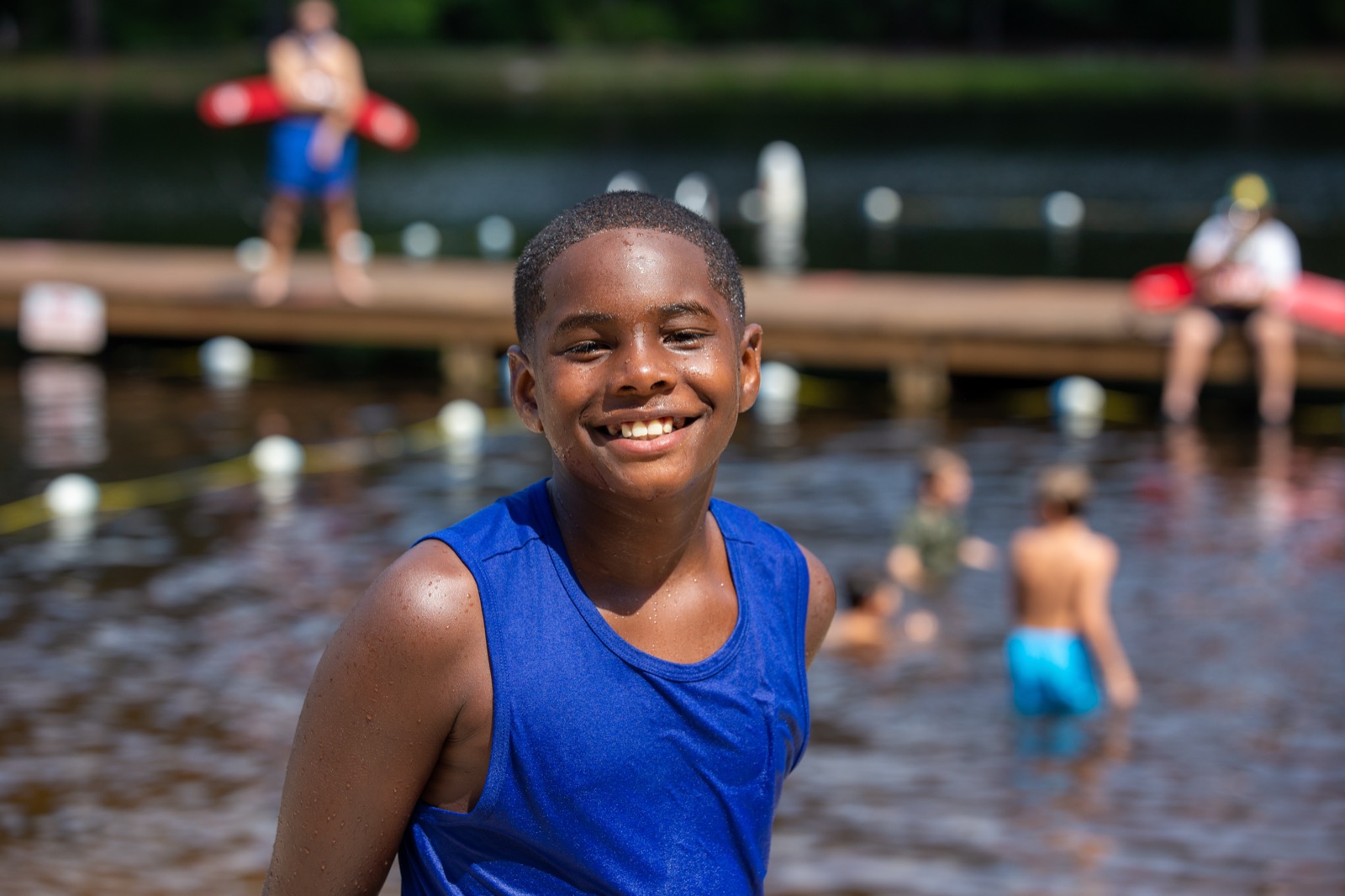 Smiling camper at the lakefront swim area, with lifeguards and swimmers in the background
