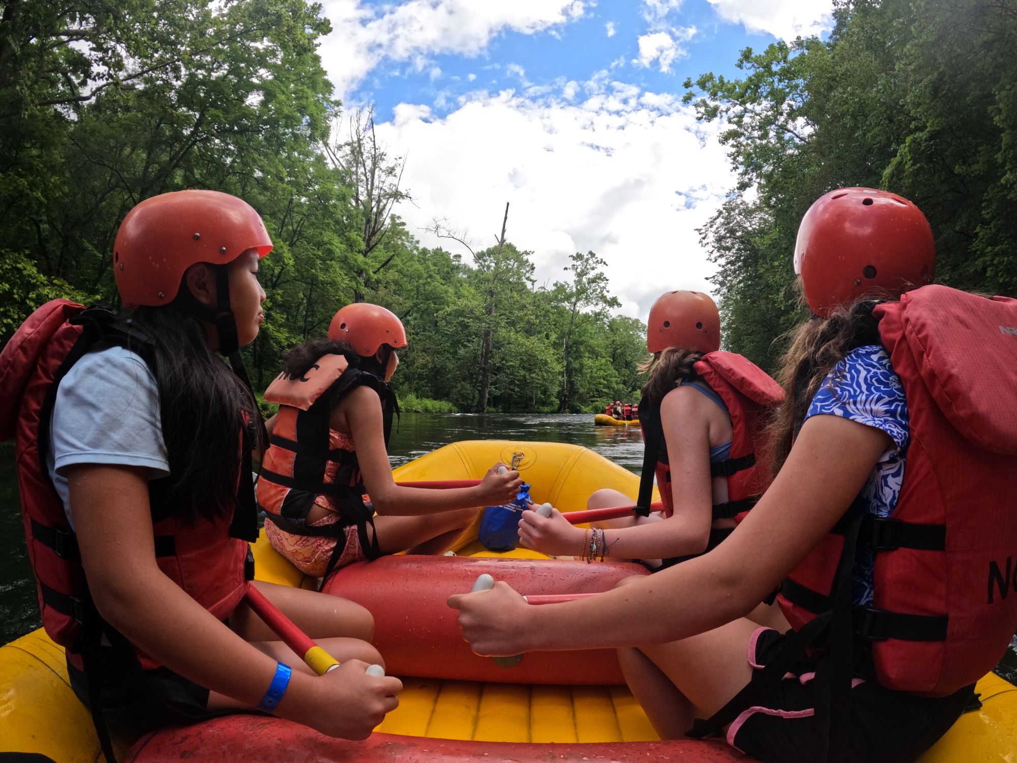 Campers in helmets and life vests paddling a raft down a tree-lined river